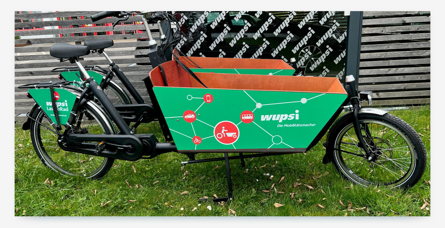 A bright green cargo bike from the "wupsi" bikesharing service. The bike's cargo box features a graphic showcasing connections to cars, buses, and trains, demonstrating a multi-modal partnership that integrates bikesharing with other forms of public and shared transport.