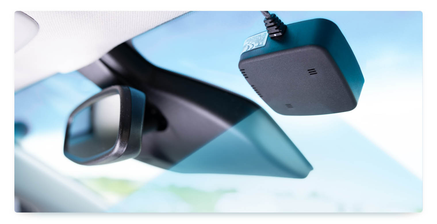 A black, compact Bosch RideCare device is attached to the inside of a car's windshield, positioned next to the rearview mirror. The device is shown with a cable running up towards the car's ceiling. The background is a clear blue sky, visible through the windshield.