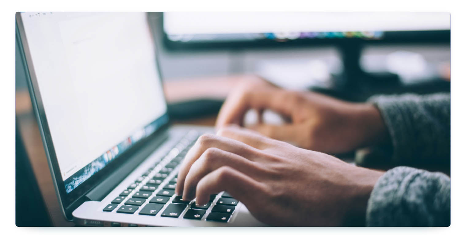 This image is a close-up shot of a person's hands typing on a laptop keyboard. The person is wearing a gray sweater, and the keyboard is illuminated by the screen. The image represents human interaction with technology, such as managing a fleet or analyzing data.