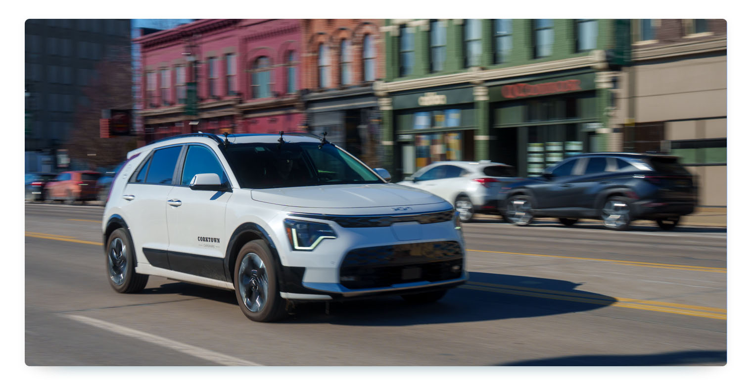 A white Kia Niro EV carsharing vehicle driving down a city street with no driver as it's being teleoperated, with colorful blurred buildings in the background. This image shows a typical shared mobility fleet vehicle in an urban environment, representing future trends that impact carsharer's proftability.