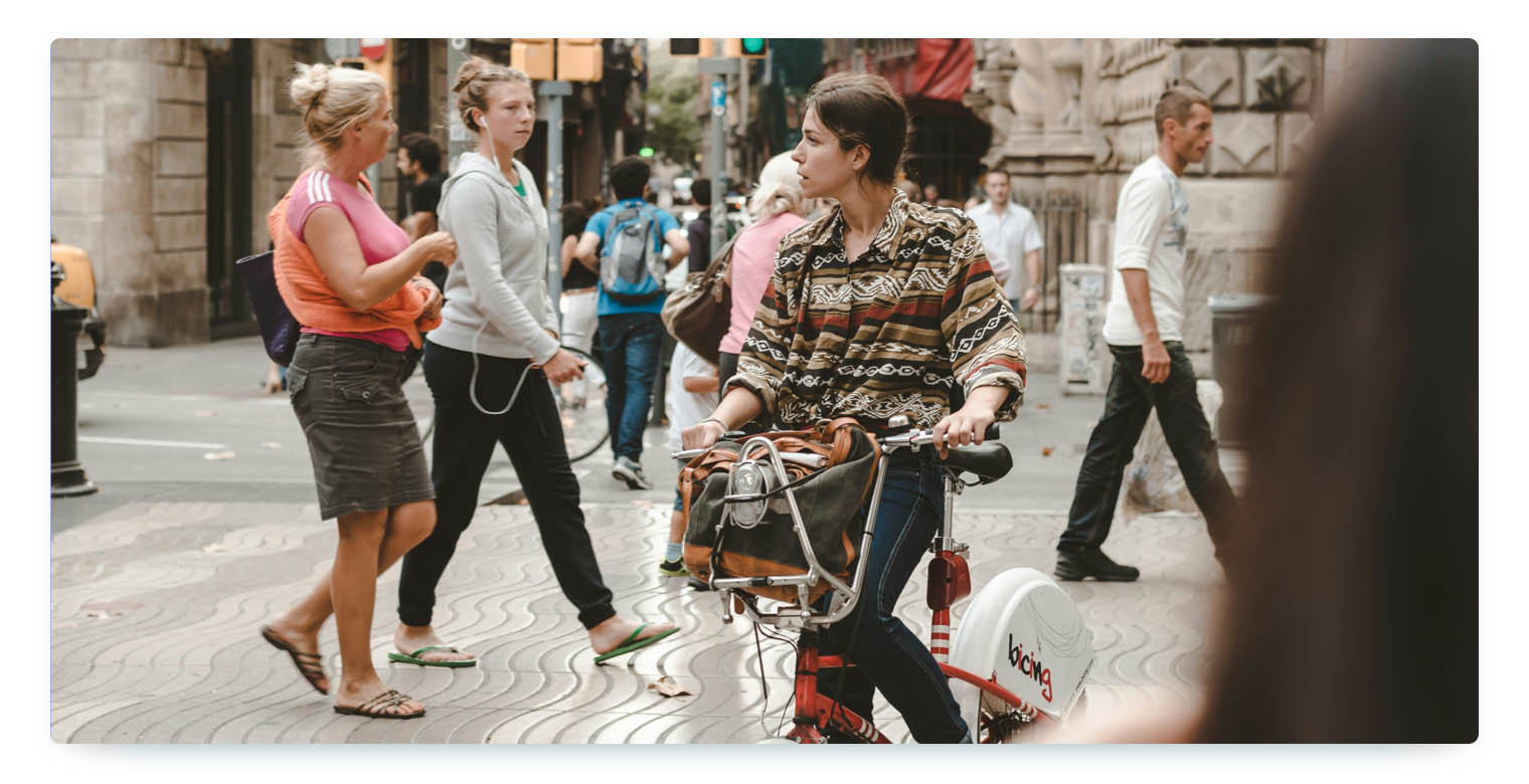 A candid street scene in a busy urban environment showing several people walking on a wavy-patterned sidewalk. In the foreground, a woman - a tourist - is riding a red bike-share bicycle with a front basket, illustrating real-world usage of shared mobility by tourists in city centers.