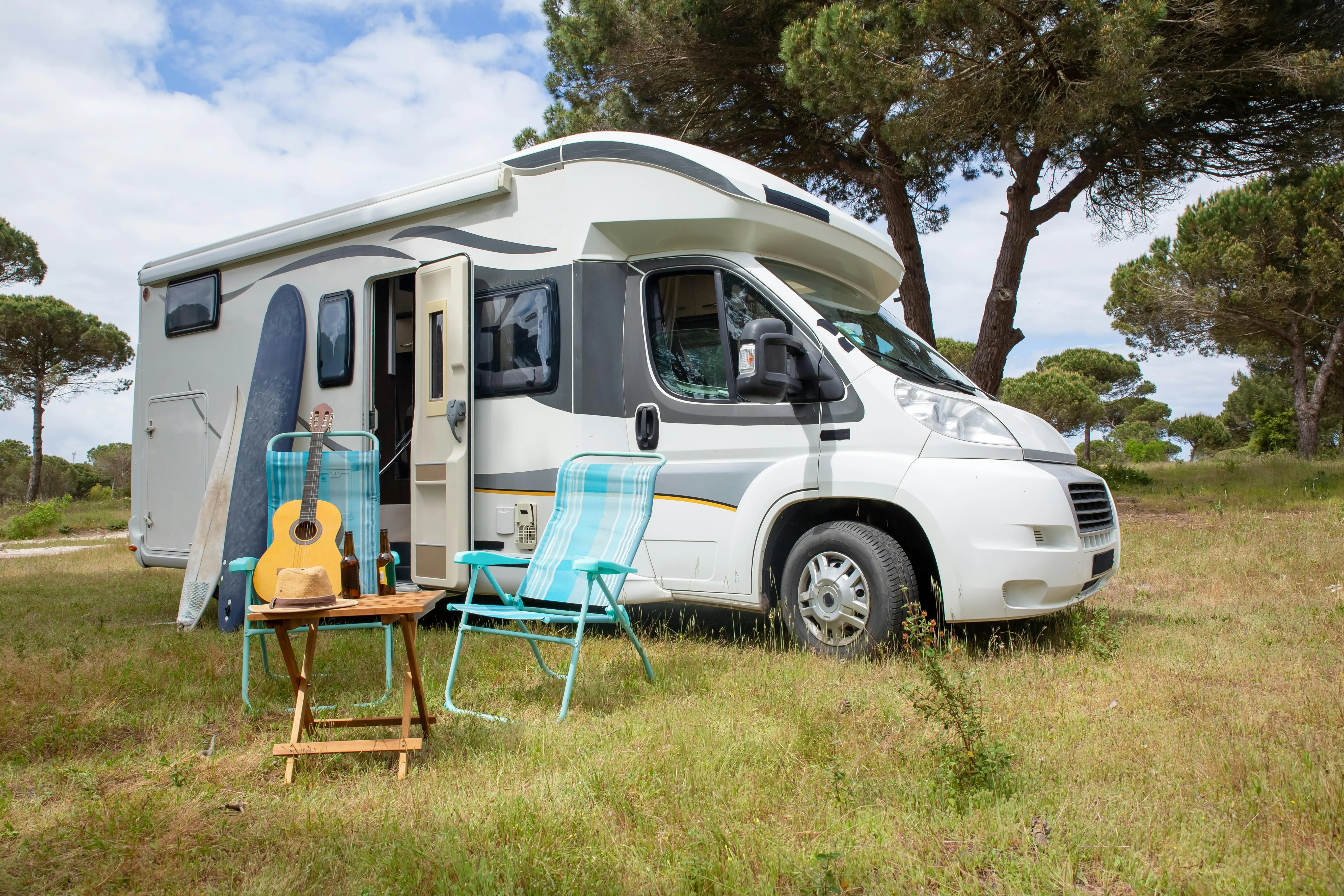 Witte camper geparkeerd op grasveld met twee blauwe stoelen, tafel met hoed en bierflesjes, een gitaar en surfplanken ernaast.