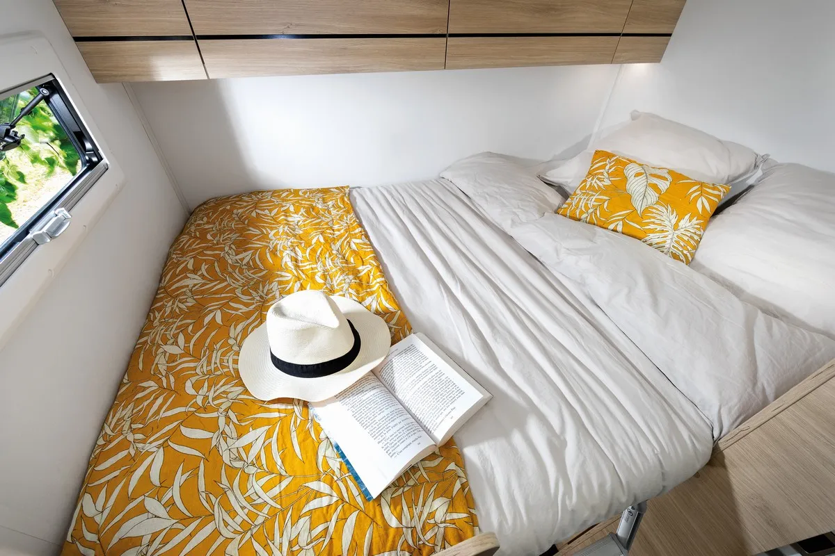 Chausson V514 camper slaapplaats met wit beddengoed, een geel kussen en een geel dekbed met bladmotief, een open boek en een witte hoed met zwarte band.