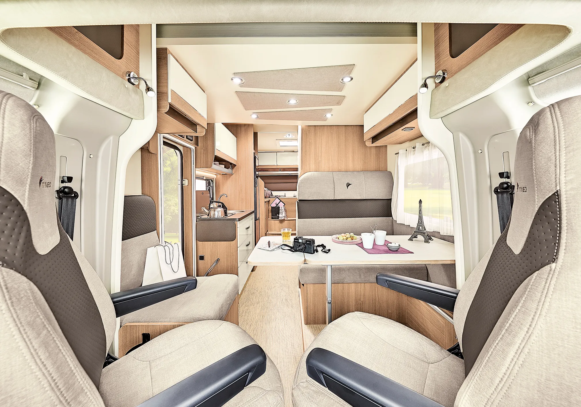 Interieur van een lichte, moderne Itineo PJ700 camper met beige stoelen, een eettafel met camera, kopjes en een miniatuur Eiffeltoren, en een compacte keuken achterin.