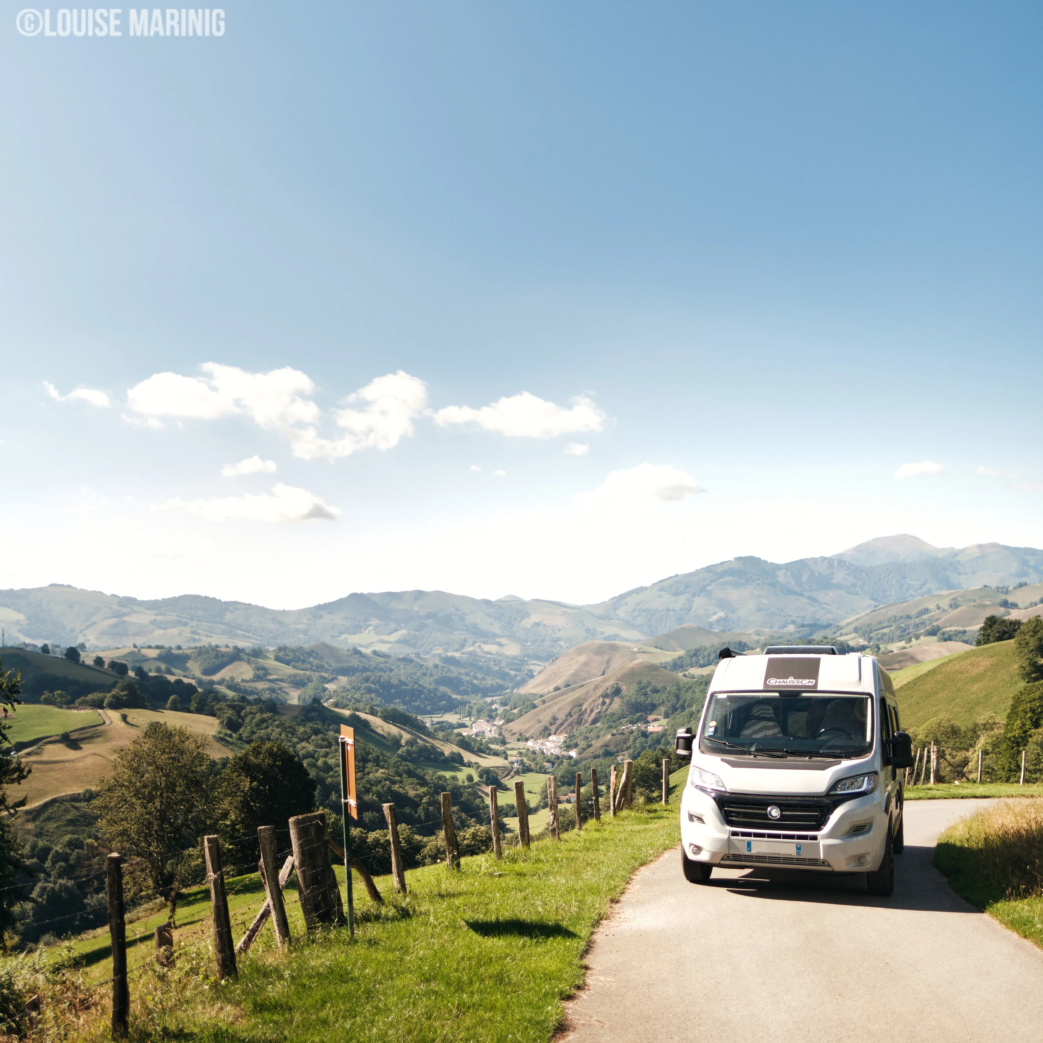 Witte Chausson camper rijdt over een smalle weg langs een houten hek met uitzicht op groene heuvels en bergen onder een bewolkte blauwe hemel.