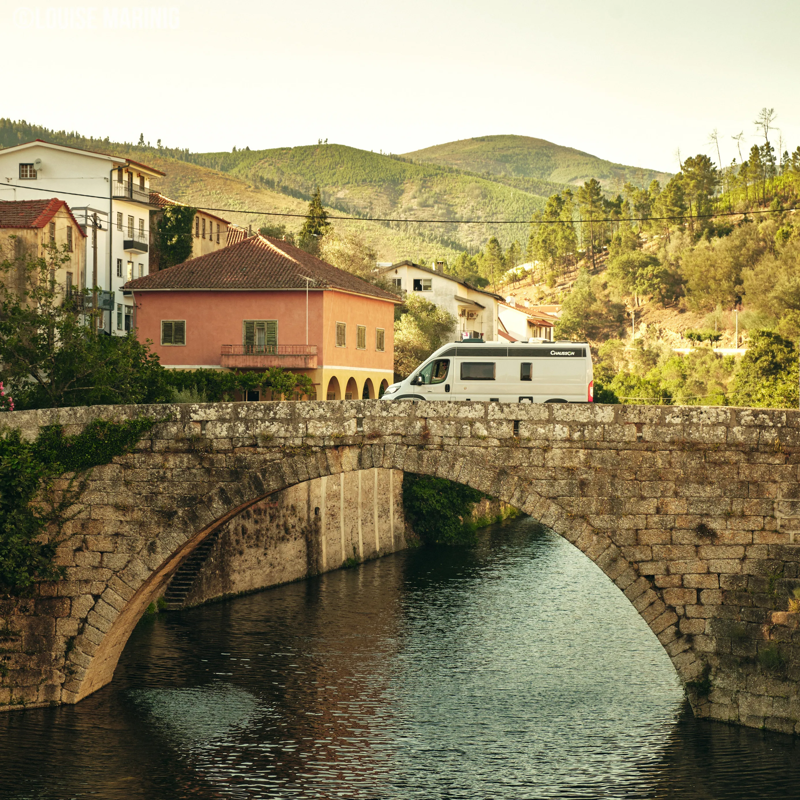 Witte camper die over een oude stenen boogbrug rijdt met huizen en groene heuvels op de achtergrond.