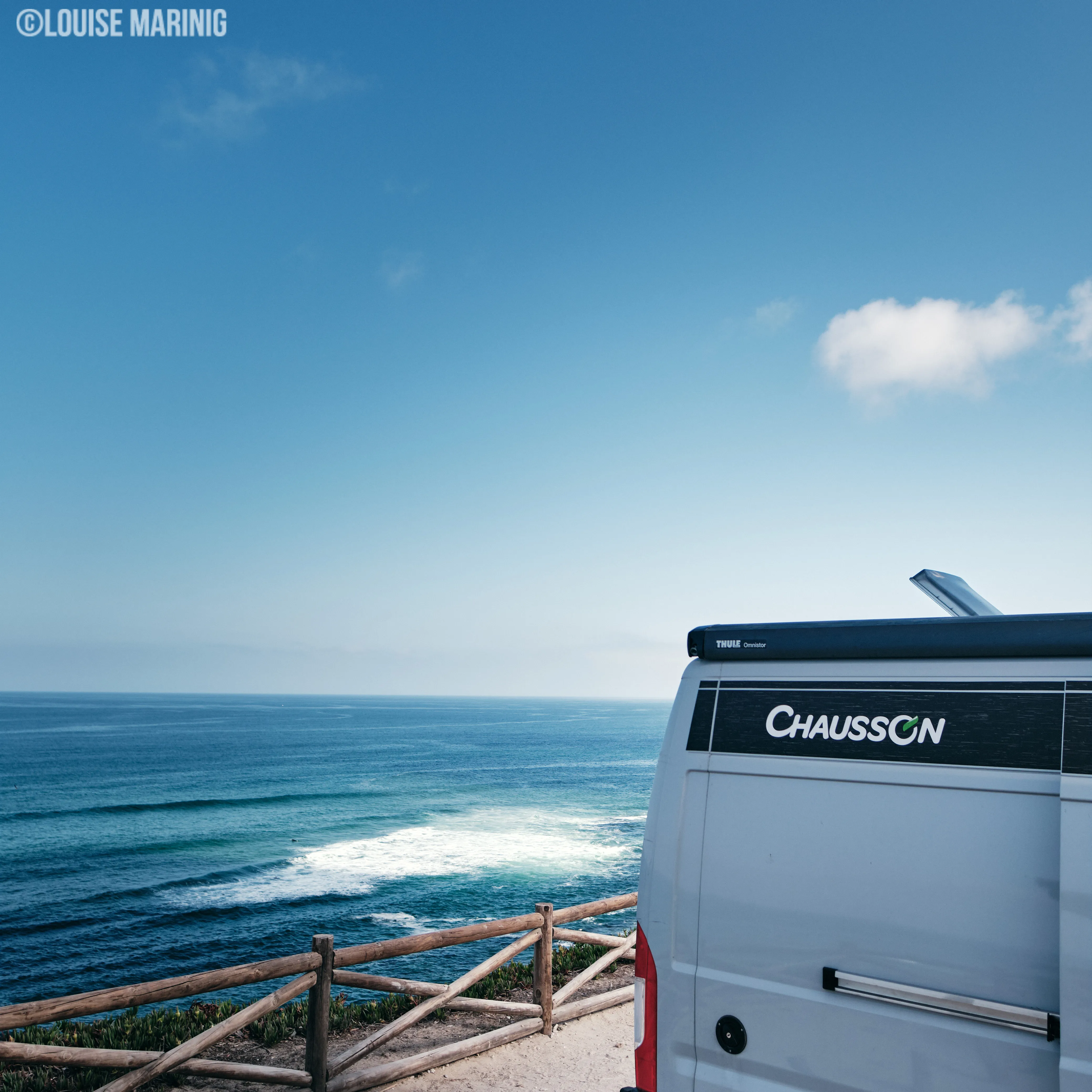 Achterkant van een witte Chausson camper geparkeerd bij een houten hek met uitzicht op een rustige oceaan en een heldere blauwe lucht.