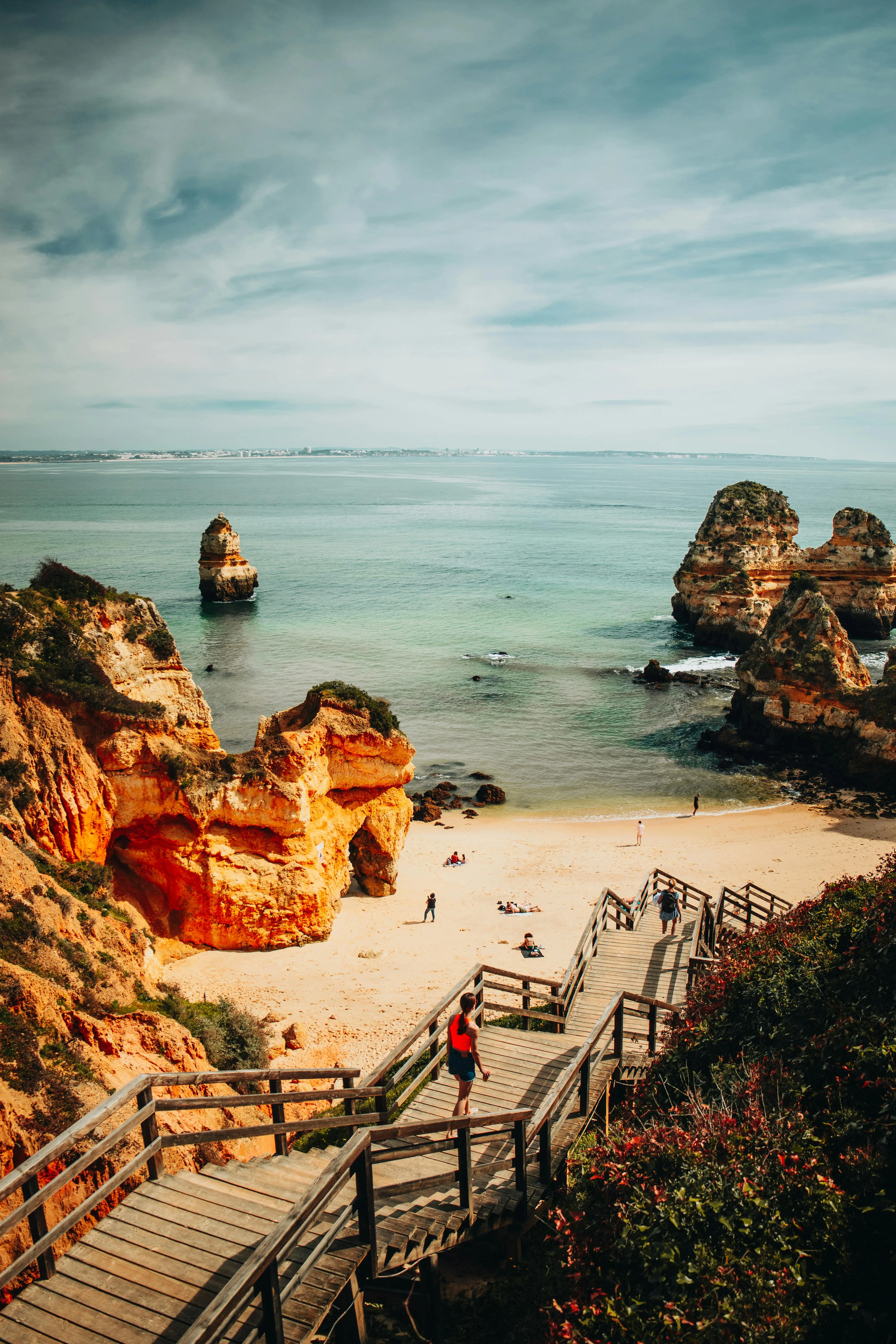 Houten trap naar een zandstrand met opvallende oranje rotsformaties en mensen die van het uitzicht genieten.