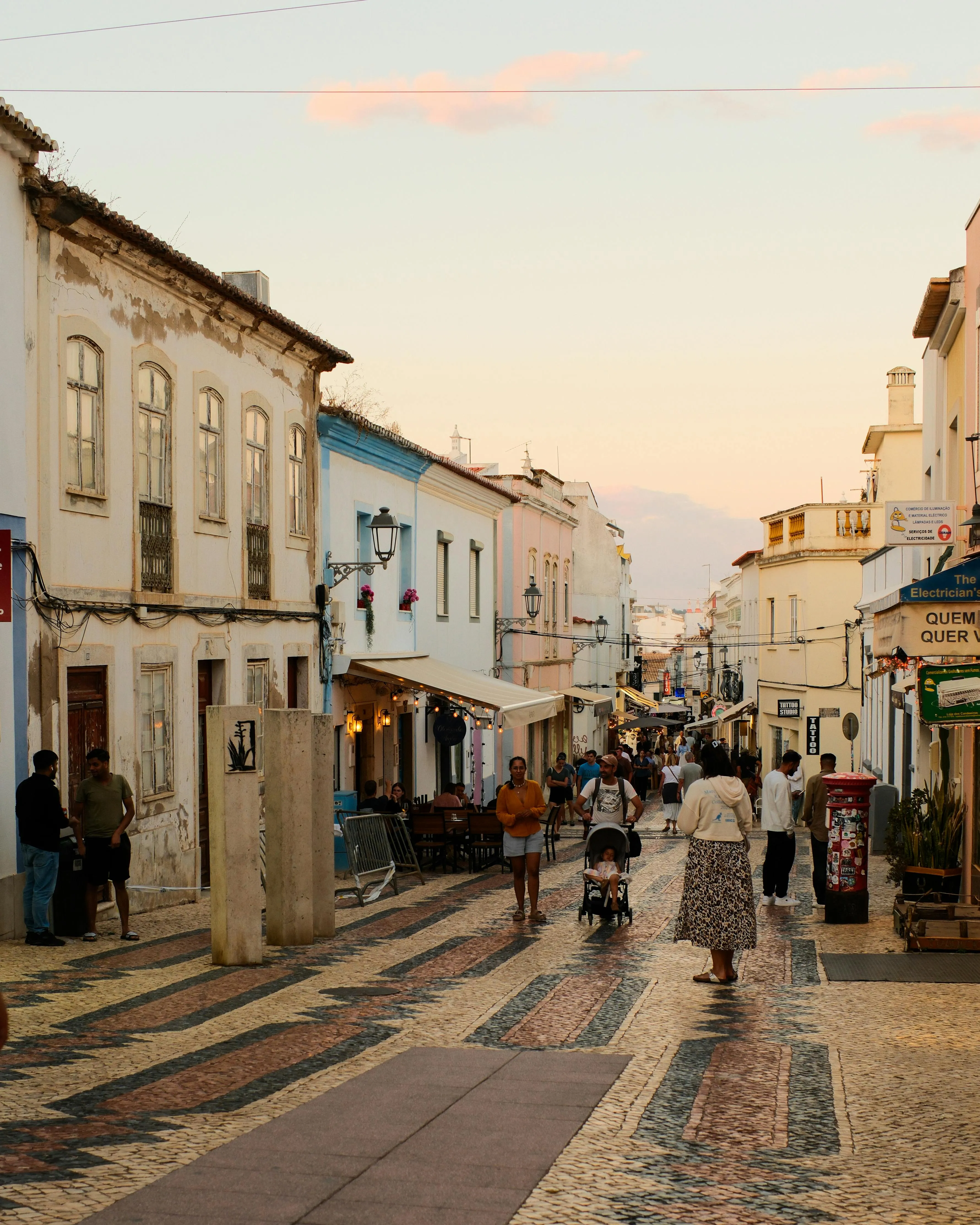Drukke straat met mensen die wandelen langs kleurrijke gebouwen en cafés tijdens zonsondergang.