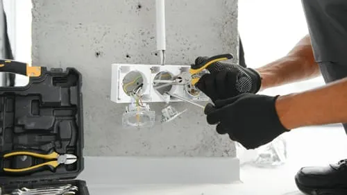 Nahaufnahme der behandschuhten Hände eines Elektrikers, der eine Steckdose an einer grauen Wand mit Werkzeugen installiert.