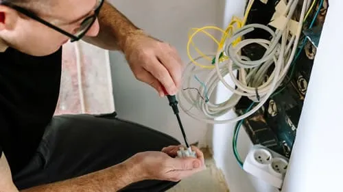 Ein Elektriker mit Brille arbeitet an einer Wandinstallation mit zahlreichen Kabeln und einer Steckdose.