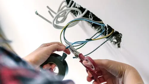 Nahaufnahme der Hände einer Person, die elektrische Kabel mit einer Schere schneidet und Isolierband hält.