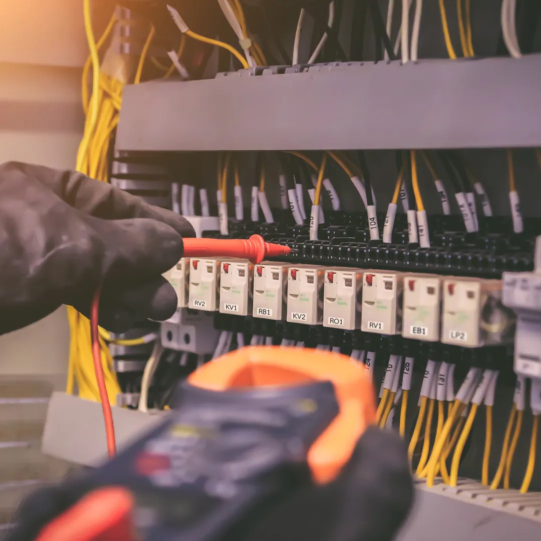 Nahaufnahme der behandschuhten Hände eines Elektrikers, der mit einem Messgerät an einem Sicherungskasten arbeitet.