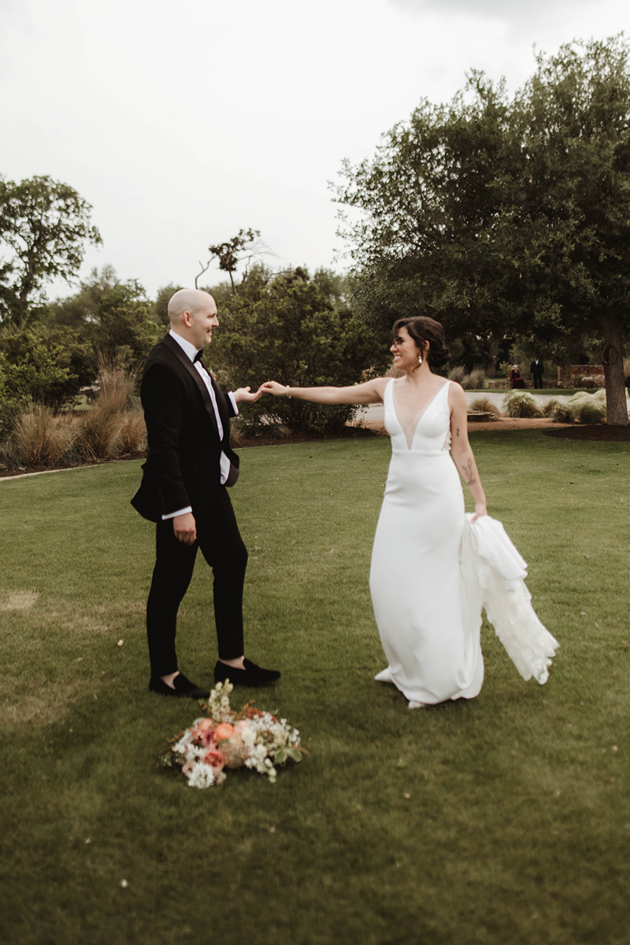 A bride and groom do a turn on a grassy field.