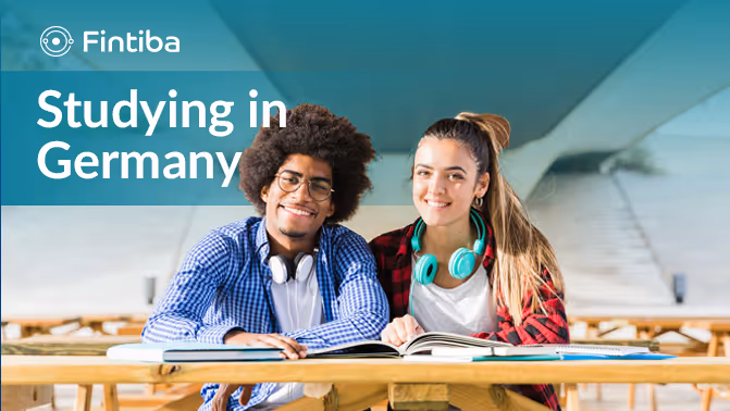 Zwei Studierende sitzen an einem Holztisch mit einem aufgeschlagenen Buch, lächelnd, unter dem Text "Studieren in Deutschland" und dem Fintiba-Logo.