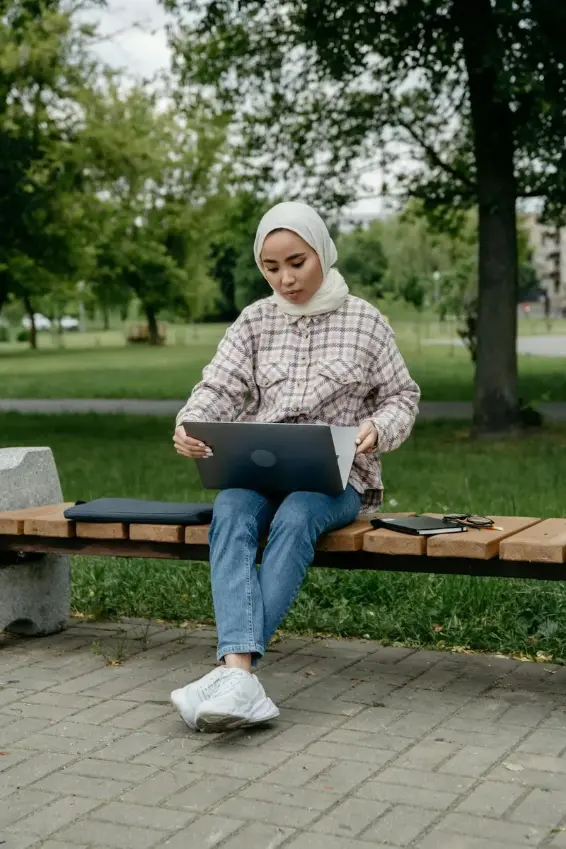 Woman wearing a hijab and checkered shirt sitting on a wooden bench in a park, using a laptop.