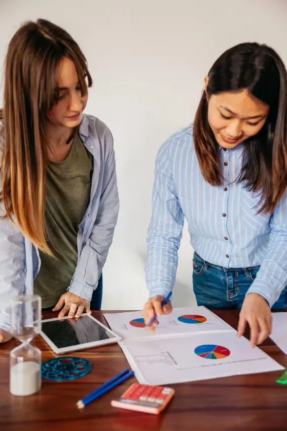 Two women reviewing colourful pie charts and graphs on paper over a wooden table with calculator, pencils, tablet, and hourglass.