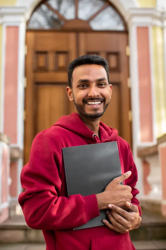 Smiling young man in a red hoodie holding a black folder, standing outside in front of a large wooden door.