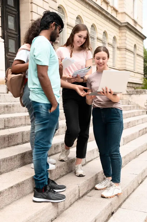Group of diverse students standing on stone steps outside a classical building, looking at a laptop and notebooks.