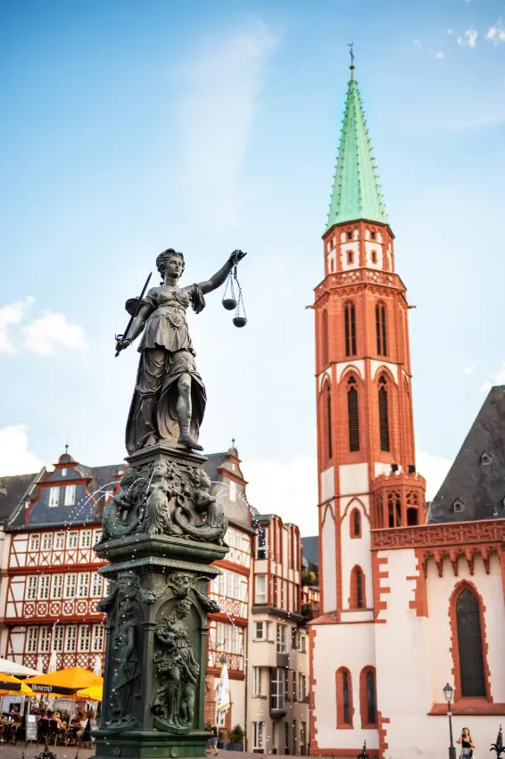 Statue of Lady Justice holding scales and sword in a German town square with traditional timber-framed buildings and a church with a green spire in the background.
