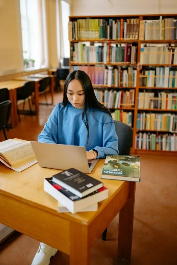 Young woman in a blue sweater working on a laptop at a wooden table in a library with bookshelves in the background.