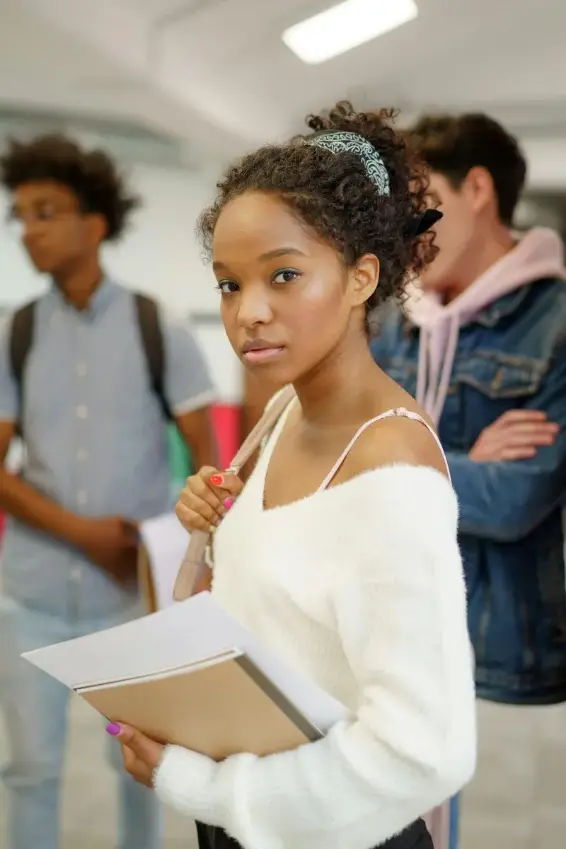 Young woman in white sweater holding folders with two students blurred in background in a school hallway.
