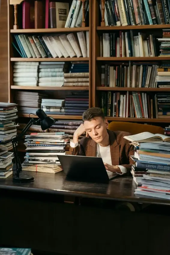 Young person sitting at a desk surrounded by stacks of books and papers, reading from a laptop with bookshelves behind.