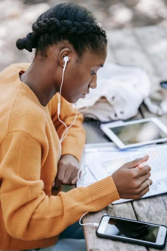 Young woman with braided hair wearing earphones and an orange sweater, reading a document at a wooden table with a smartphone and tablet nearby.