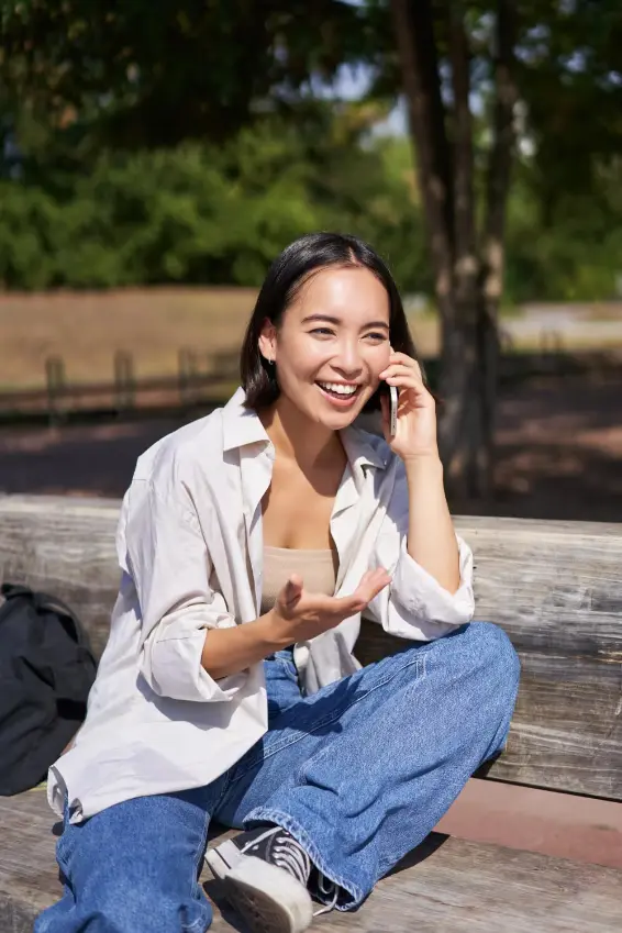 Smiling young woman sitting on a wooden bench outdoors, talking on a mobile phone.