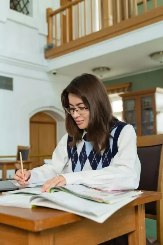 Young woman with glasses studying and writing at a wooden desk in a bright, spacious room.