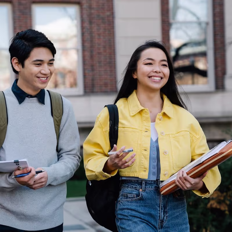 Two smiling students walking outside a building, one wearing a yellow jacket and carrying folders, the other in a grey sweater with a backpack. Discussing financial proof for german visa.