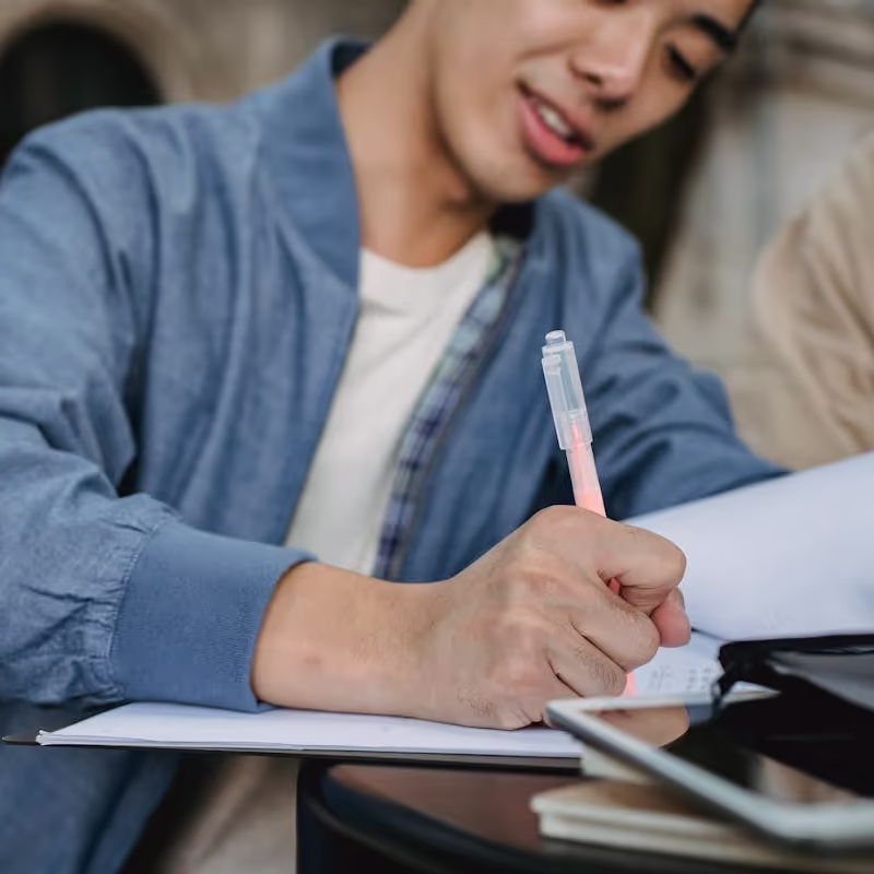 Young man in blue jacket writing with a pen in a notebook at a table, learning about blocked accounts for german visa