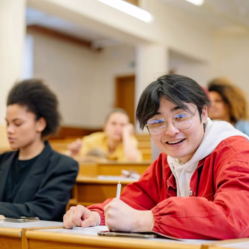 Smiling student in glasses and red jacket writing on paper in a classroom with other students.