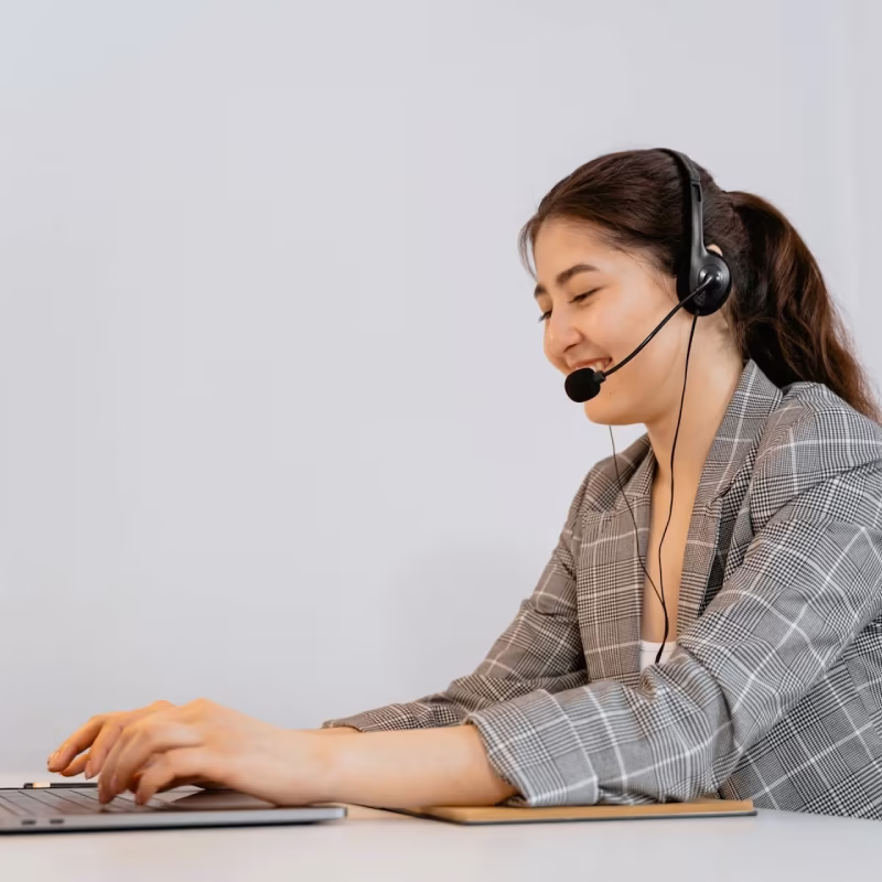 Smiling woman wearing a headset and checked blazer typing on a laptop at a white desk.