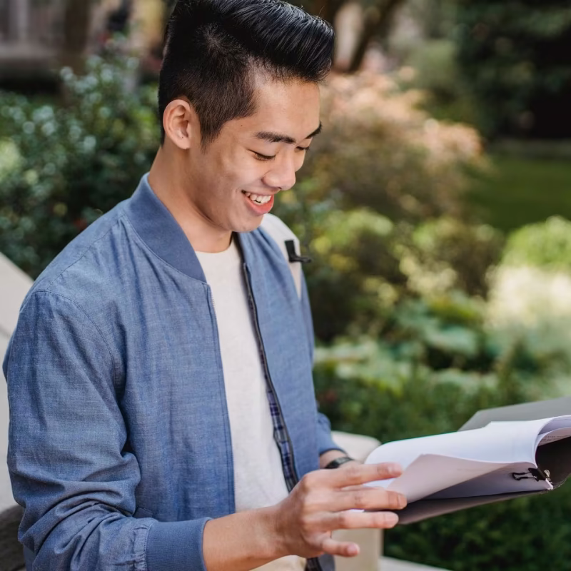 Young man smiling and reading an open book outdoors with greenery in the background.