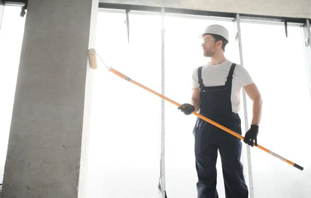Maler in Arbeitskleidung und Schutzhelm, der eine Wand mit einer Farbrolle an einem langen Stiel streicht.