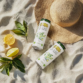 Two Perico Limojito energy drinks placed on sunlit beach sand next to sliced lemon, green leaves, and a woven sunhat, showcasing tropical travel vibes. 