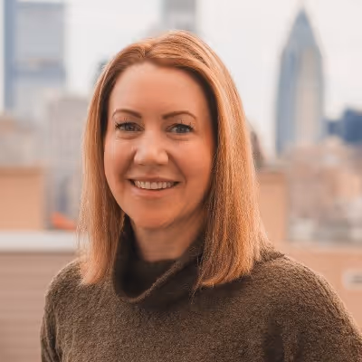 Smiling woman with straight shoulder-length light brown hair wearing a brown sweater with a city skyline blurred in the background.