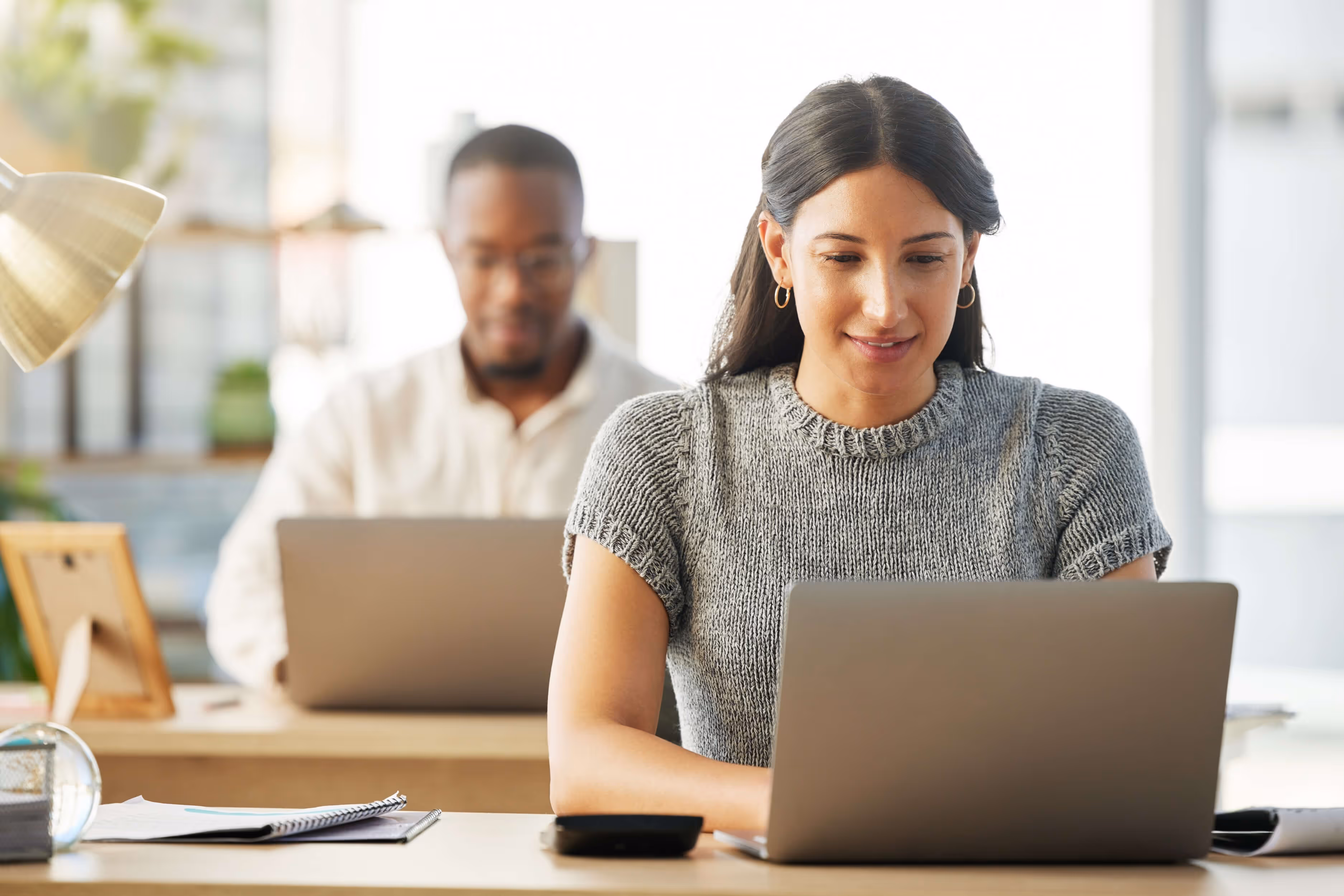 Two colleagues working on laptops at desks in a bright office space.