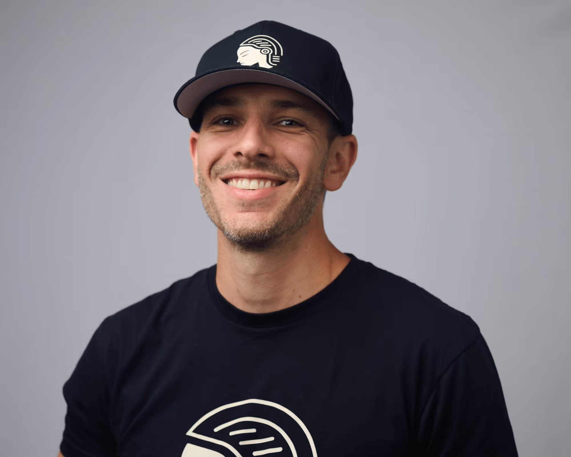 Smiling man wearing a black cap and black t-shirt with a white logo against a plain gray background.