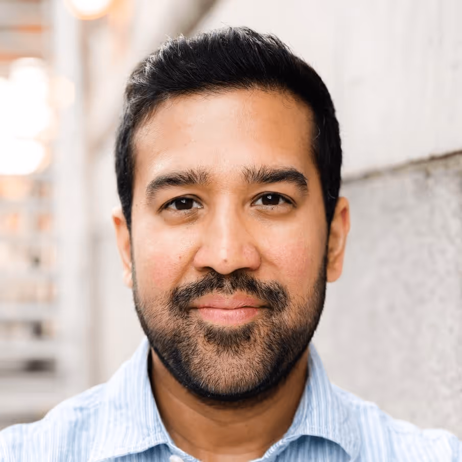 Smiling man with dark hair and beard wearing a light blue striped shirt, standing against a blurred urban background.