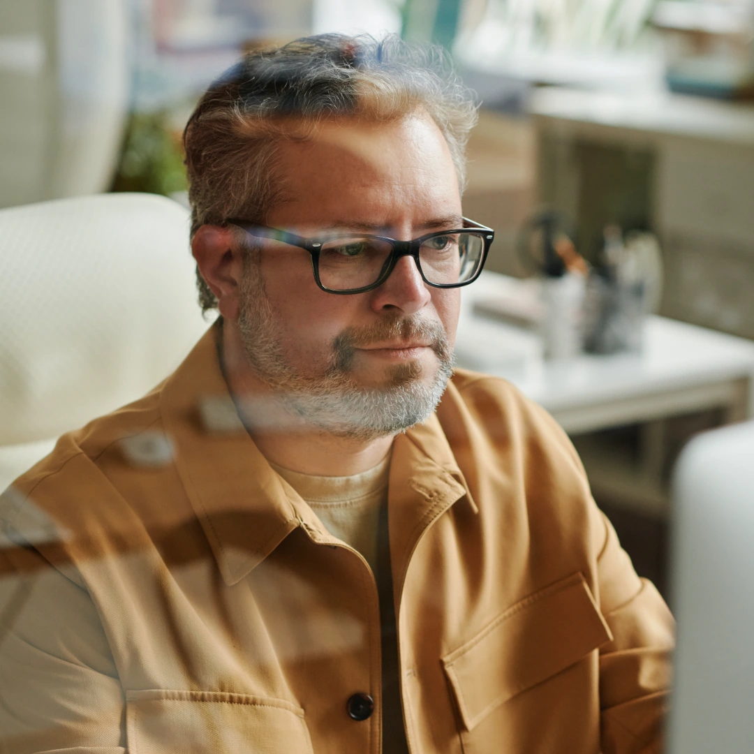 Middle-aged man with glasses and a beard wearing a brown jacket, focused on a computer screen indoors.