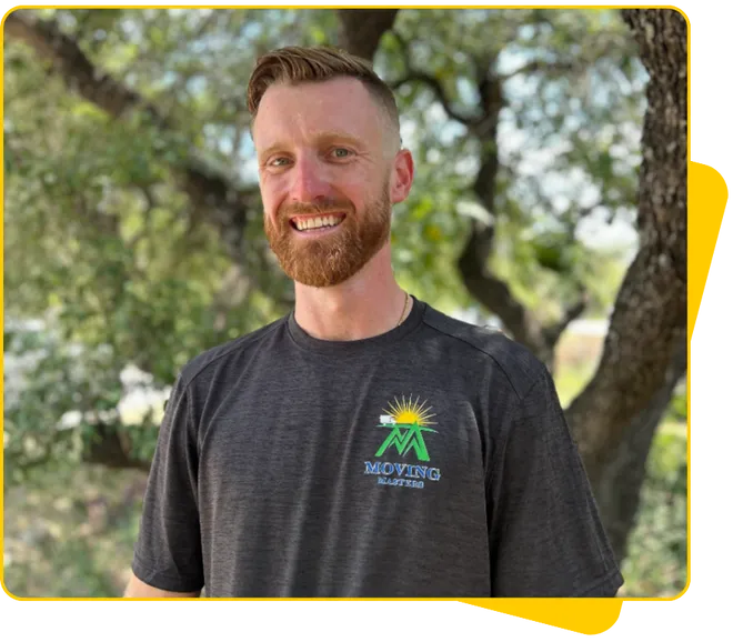 Smiling man with short light brown hair and beard wearing a dark gray Moving Masters t-shirt standing outdoors with trees in the background.