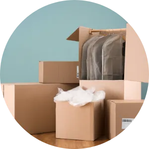 A group of cardboard moving boxes, one open with plastic garment covers hanging inside, another smaller open box with white packing material spilling out, set on a wooden floor against a blue wall.