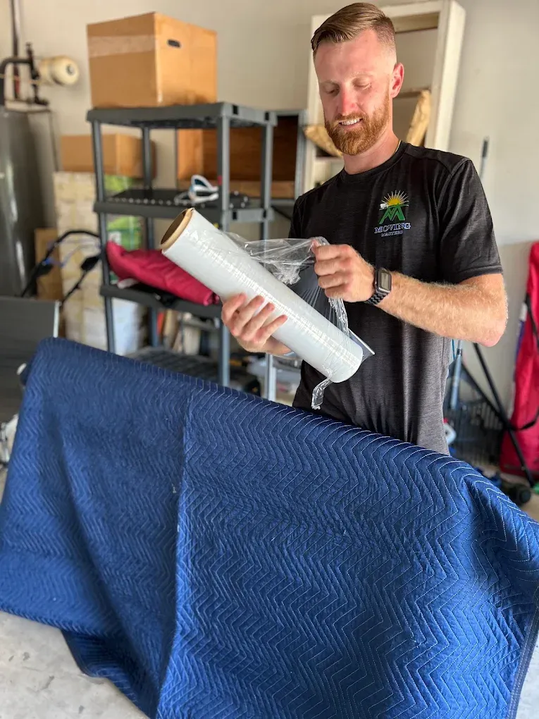 A man with a beard is preparing packing material by unrolling clear plastic wrap in a storage or workshop area with shelves and various items in the background, and a blue padded moving blanket in the foreground.