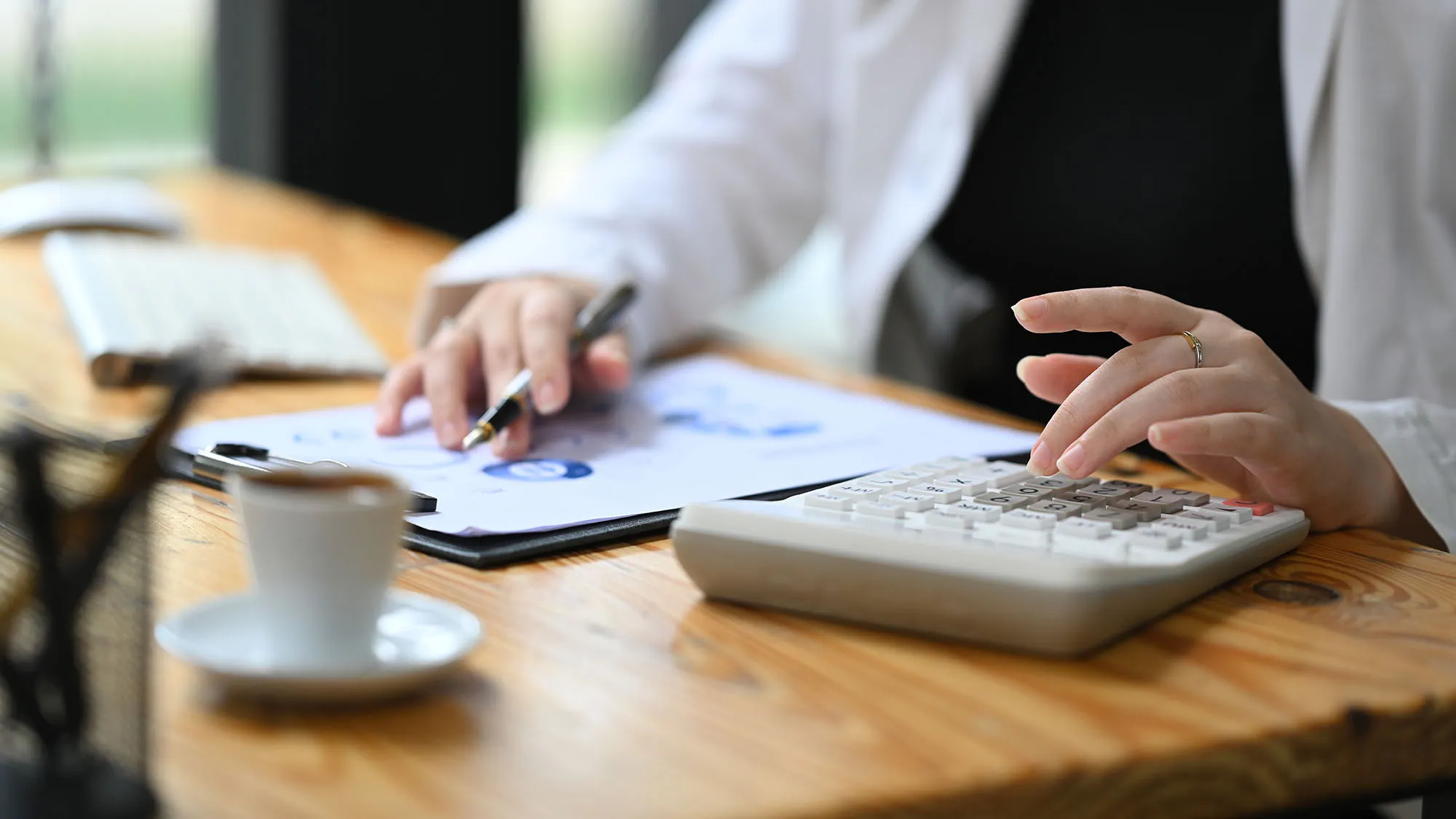Close-up of a person working at a desk, using a calculator with one hand and holding a pen over financial documents with charts.
