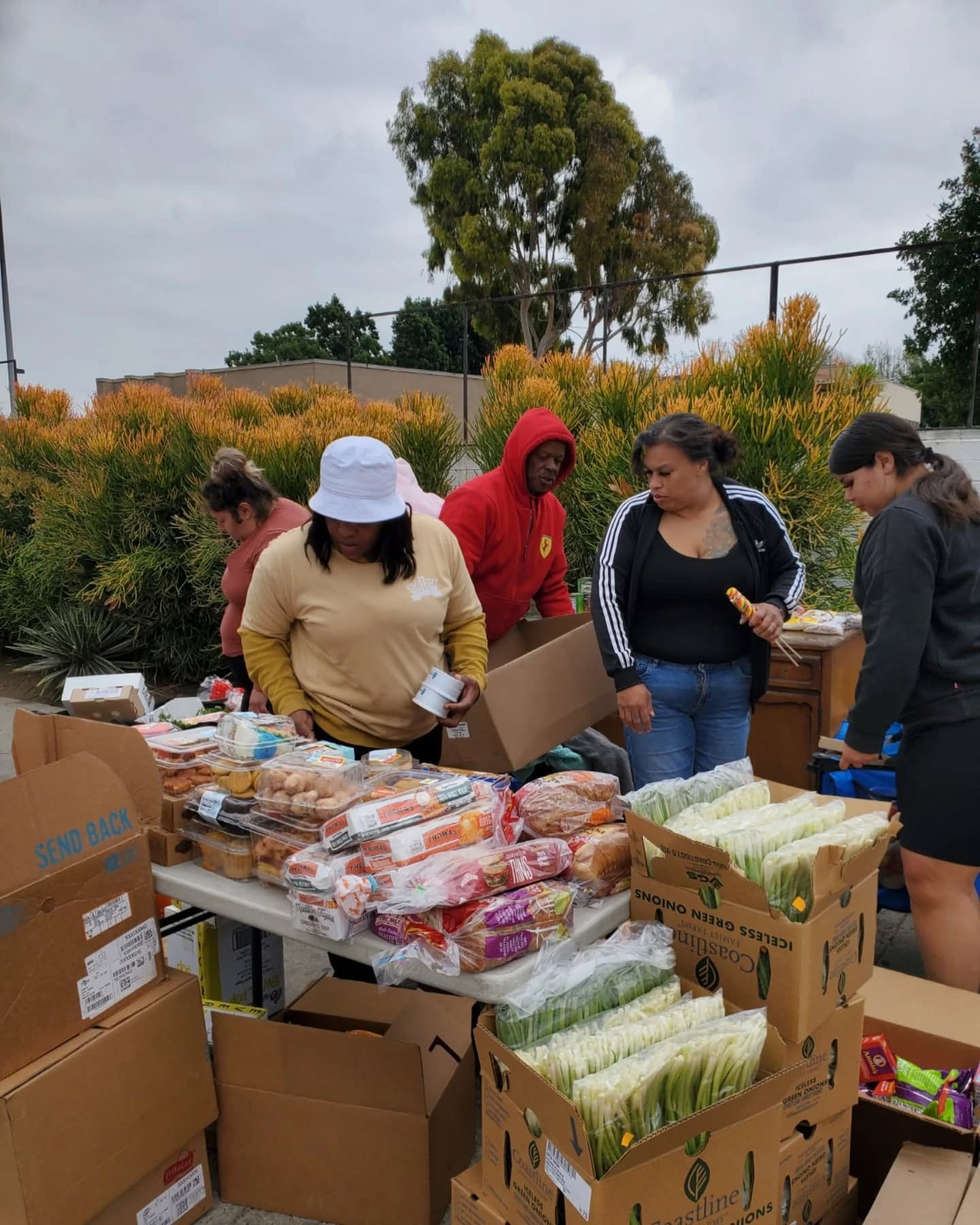 Community members gathered around tables filled with packaged food items, organizing and distributing groceries outdoors on a cloudy day surrounded by greenery.