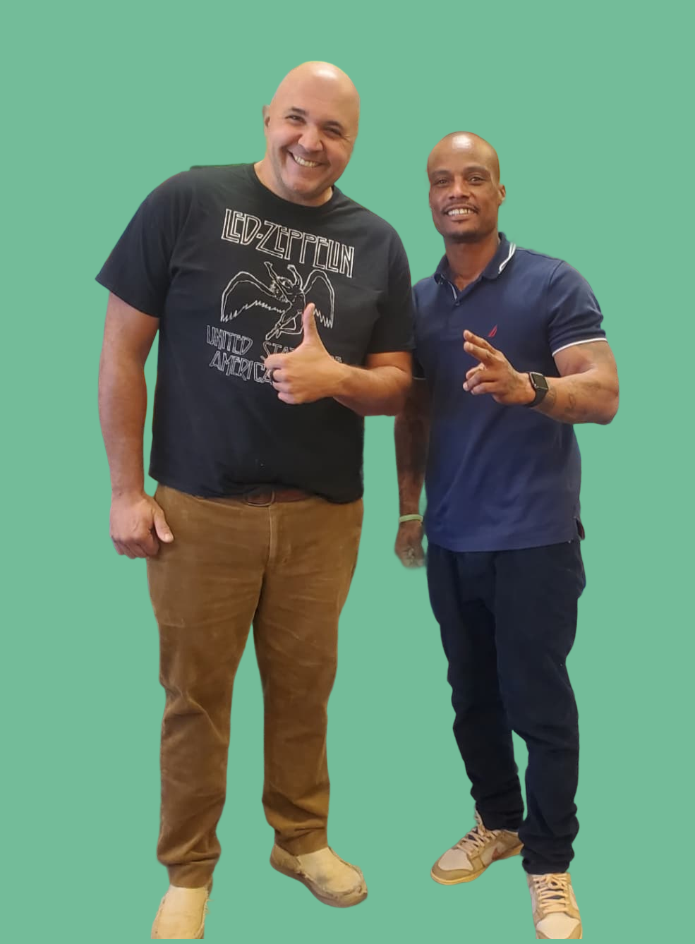Two men standing and smiling in front of a green background, one giving a thumbs up and the other flashing a peace sign, symbolizing community and camaraderie.