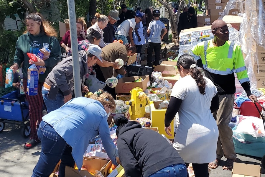Volunteers and community members gather around tables full of grocery items at an outdoor food distribution event.