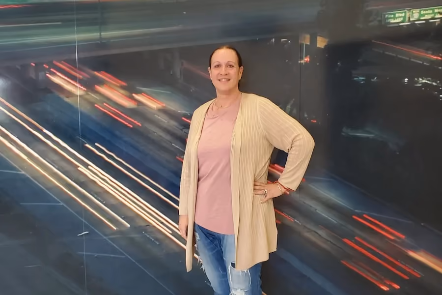 Yana Shmuliver, founder of K.A.U.S.E., in a long beige cardigan and jeans poses in front of a mural of highway light trails.